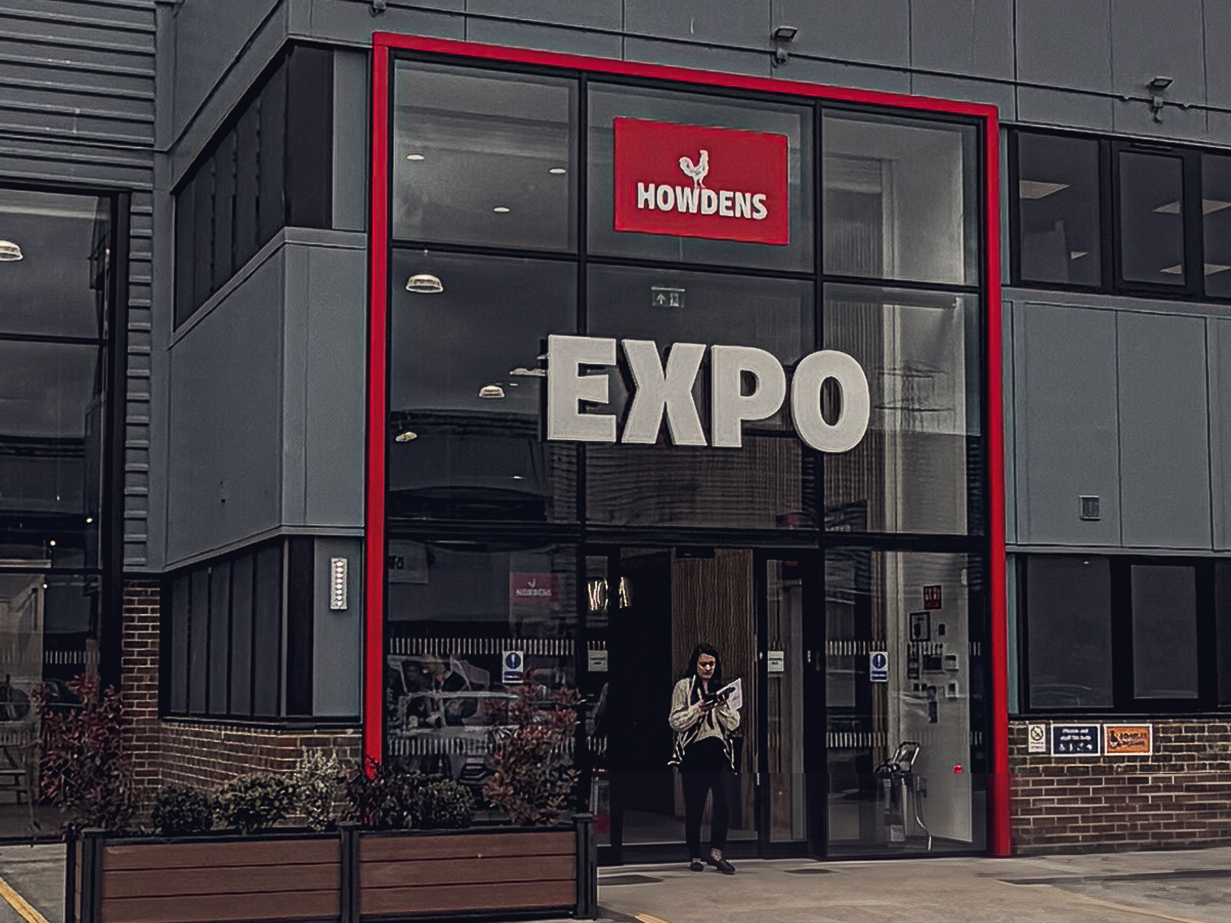 The red-framed glazed entrance beacon of Howdens Expo in Watford, set into grey profile metal cladding above a brick plinth, by we are _
