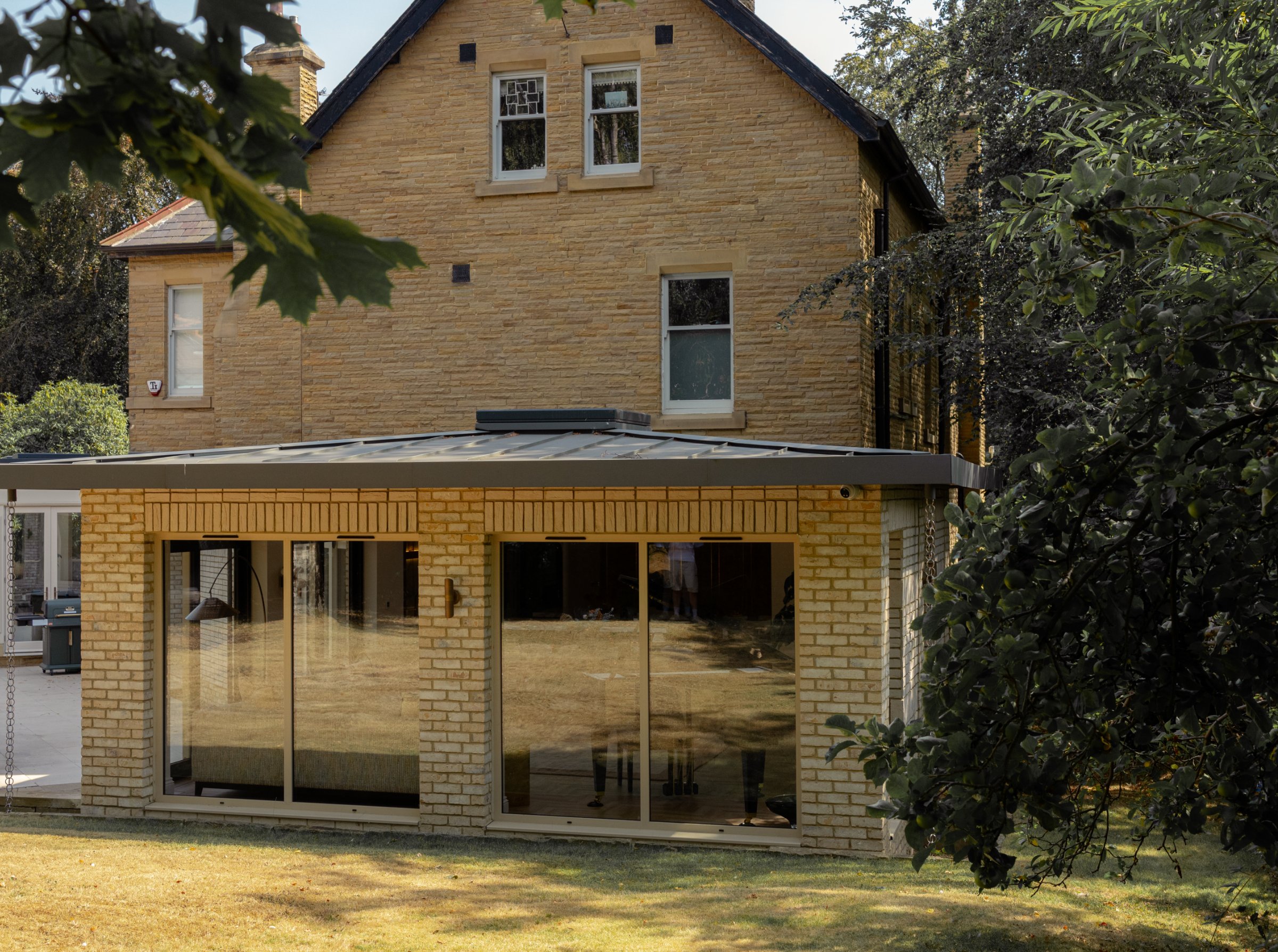 Garden pavilion extension behind a sandstone Victorian villa in Roundhay, Leeds, by we are _, with buff brick piers, a low metal roof and full-height sliding glazing onto the lawn